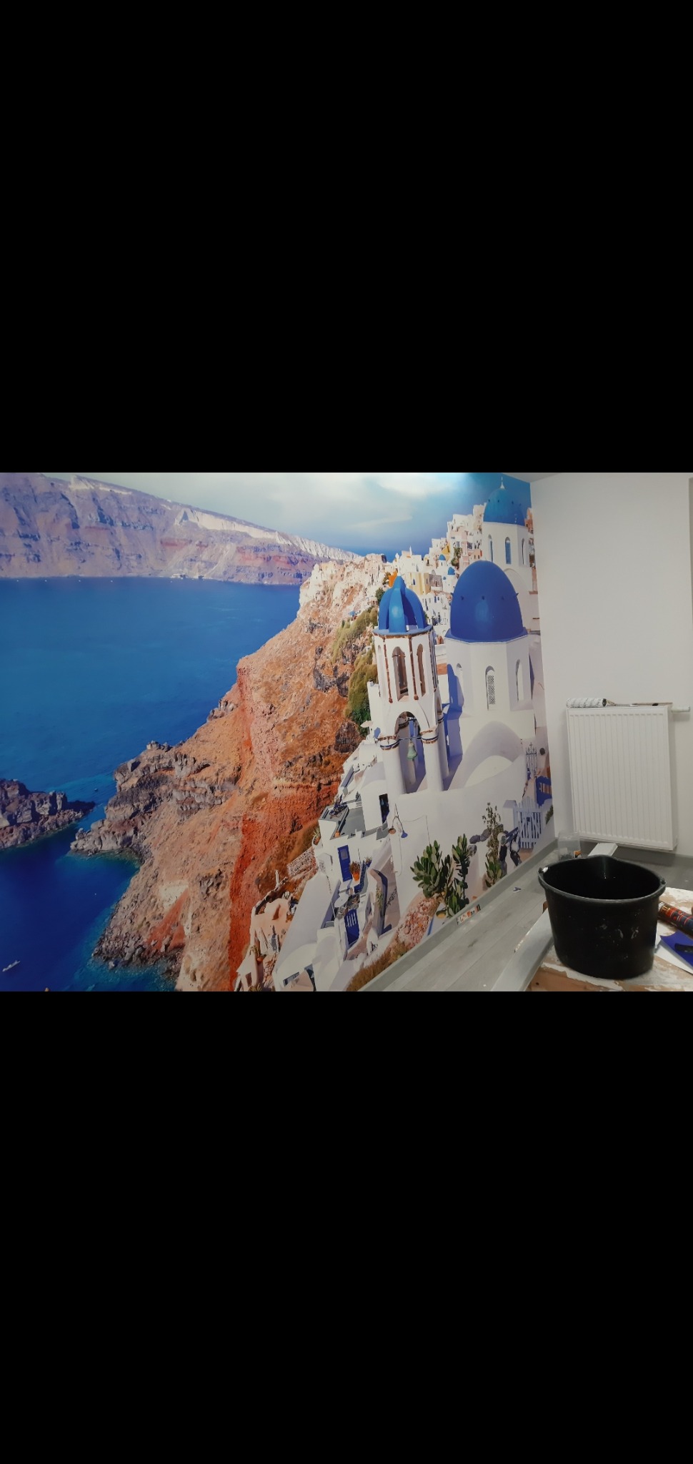 Naklejona tapeta z motywem Santorini na ścianie w Bydgoszczy, widoczny czarny kubeł i grzejnik w tle.