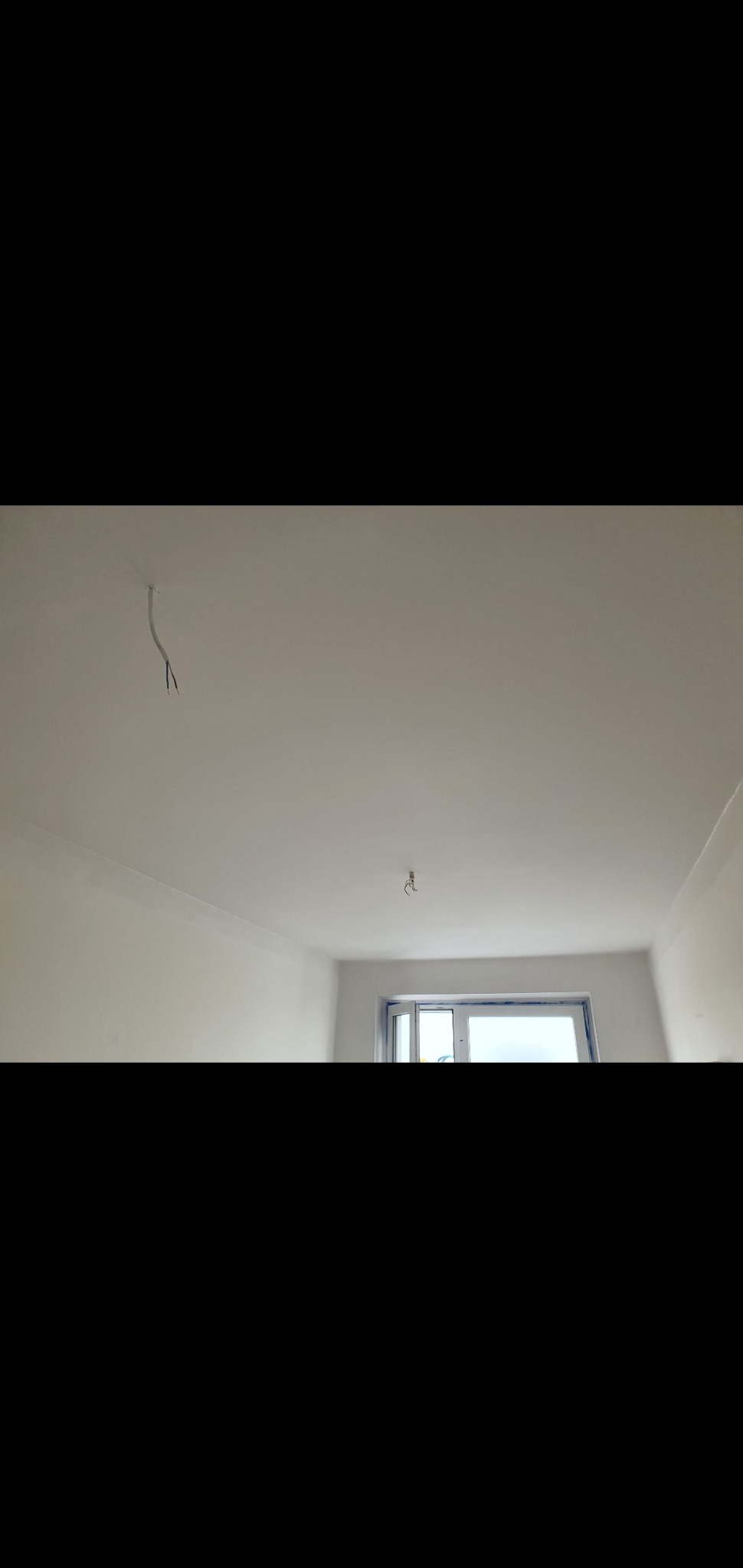 Pomalowane na biało wnętrze pokoju z wystającymi przewodami z sufitu i oknem w tle.