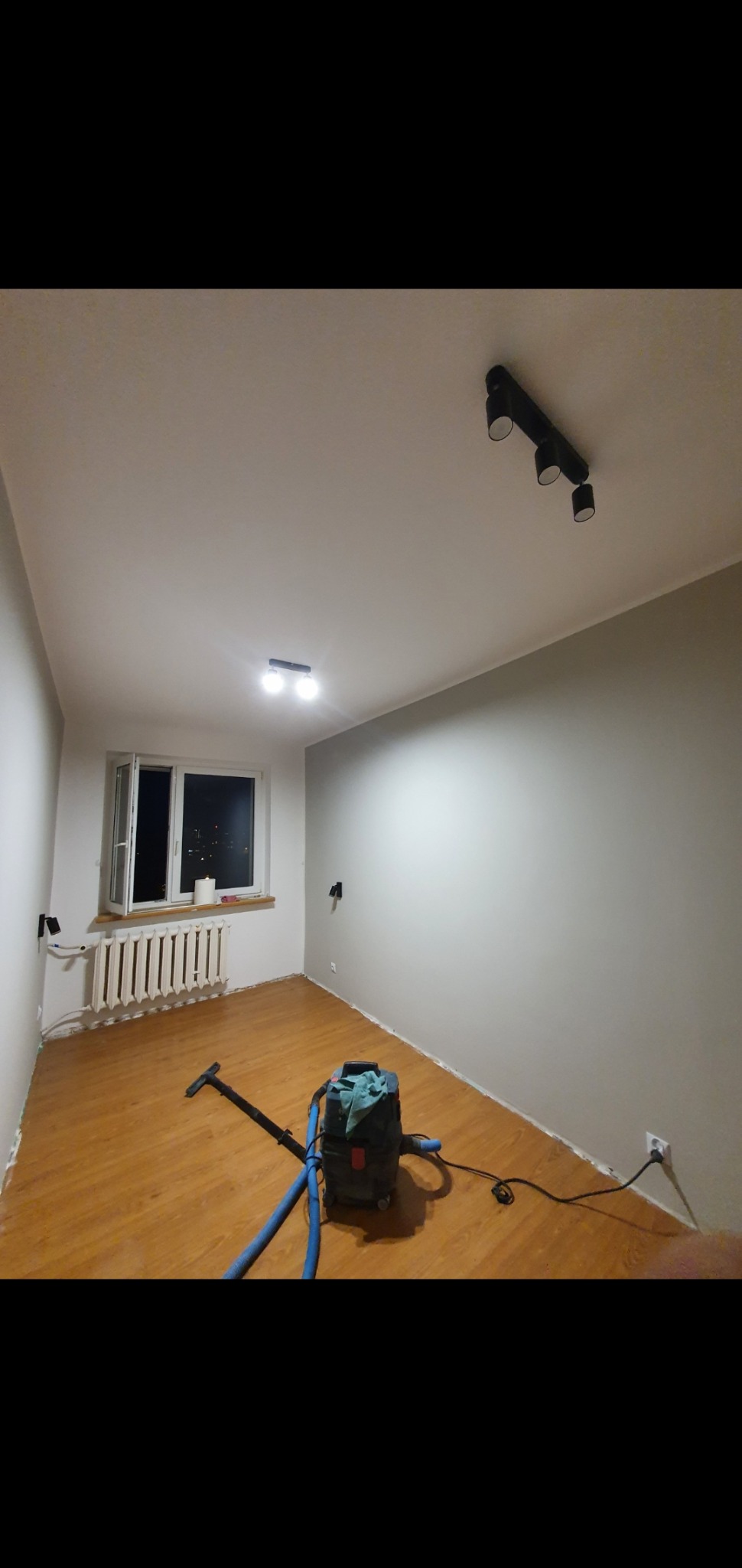 Pokój po malowaniu z drewnianą podłogą, widoczny odkurzacz przemysłowy i grzejnik pod oknem z widokiem na nocne miasto.