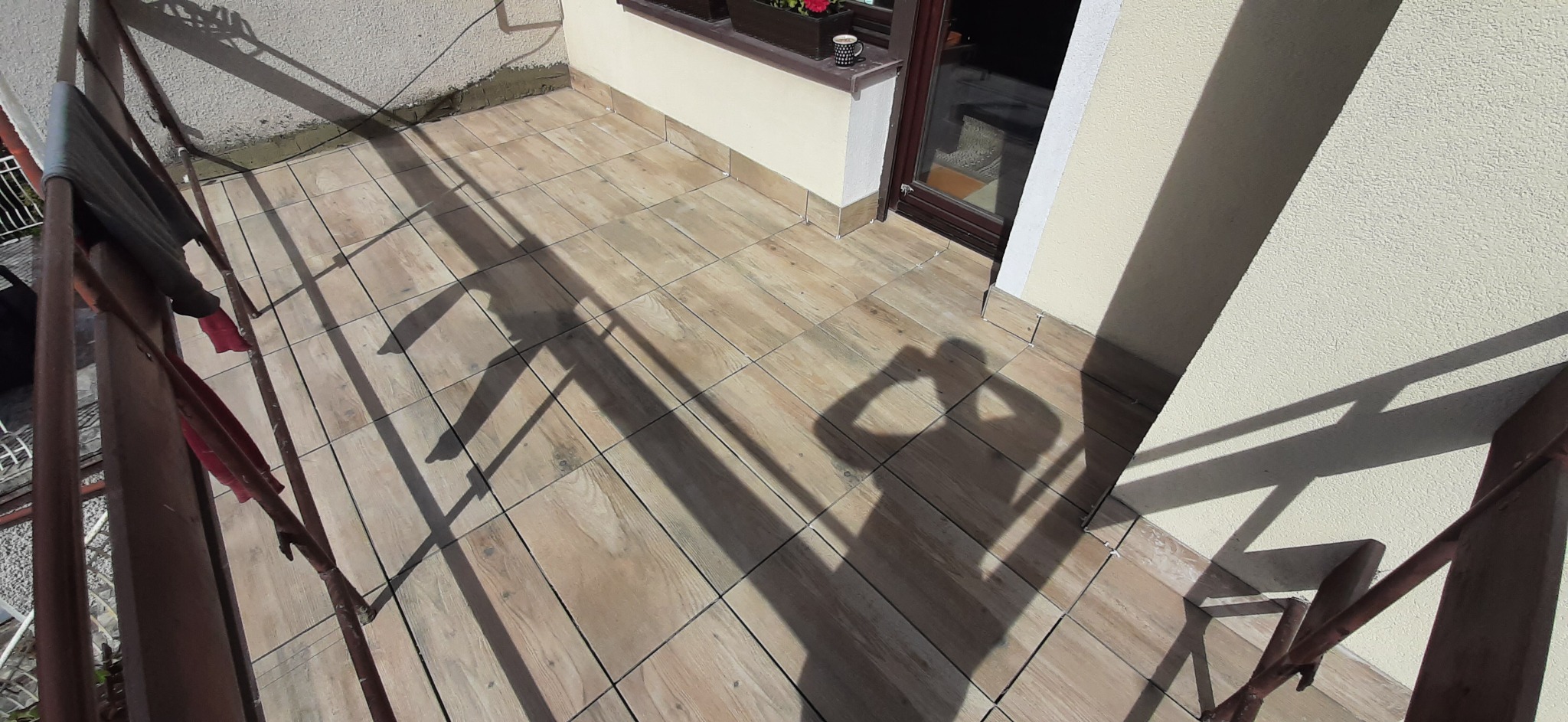 Wyłożony płytkami balkon z drewnopodobnym wzorem, widoczny cień osoby robiącej zdjęcie, brązowa balustrada i fragment ściany z oknem, na parapecie stoi kubek.