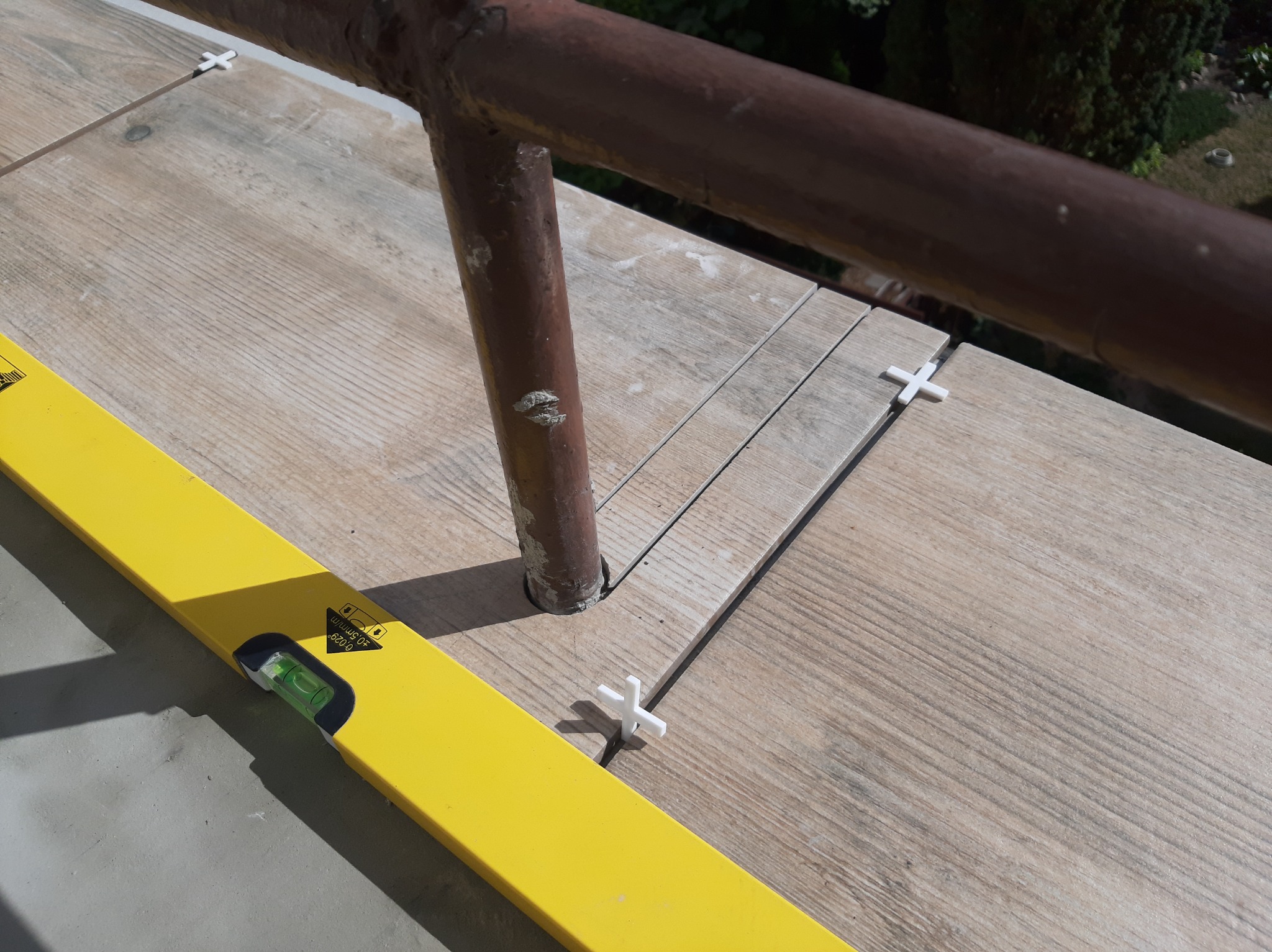 Układanie płytek imitujących drewno na balkonie, poziomnica i krzyżyki dystansowe w użyciu, widoczna metalowa balustrada.