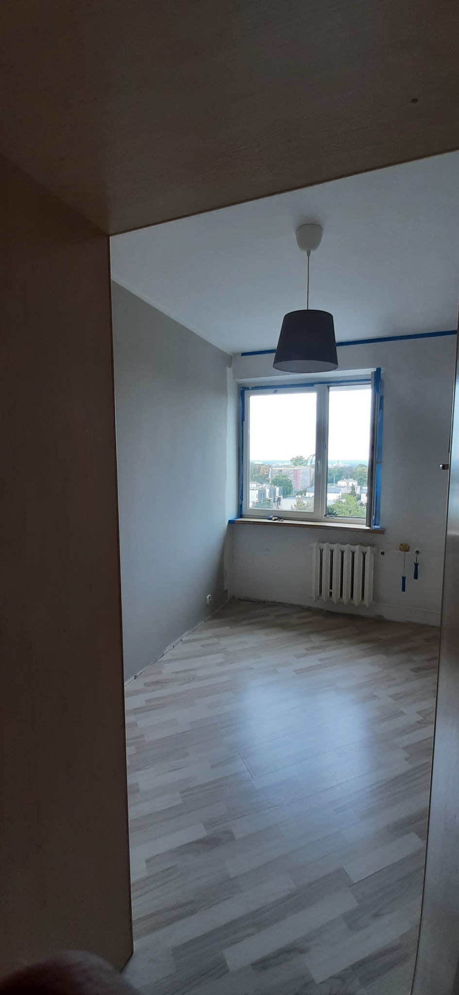 Pokój po malowaniu ścian na szaro, okno zabezpieczone taśmą malarską, widoczny grzejnik i lampa sufitowa.
