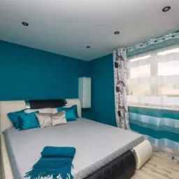 Turkusowa sypialnia z łóżkiem, zasłonami w wieże Eiffla i drewnianą podłogą, oświetlona punktowymi lampami sufitowymi.