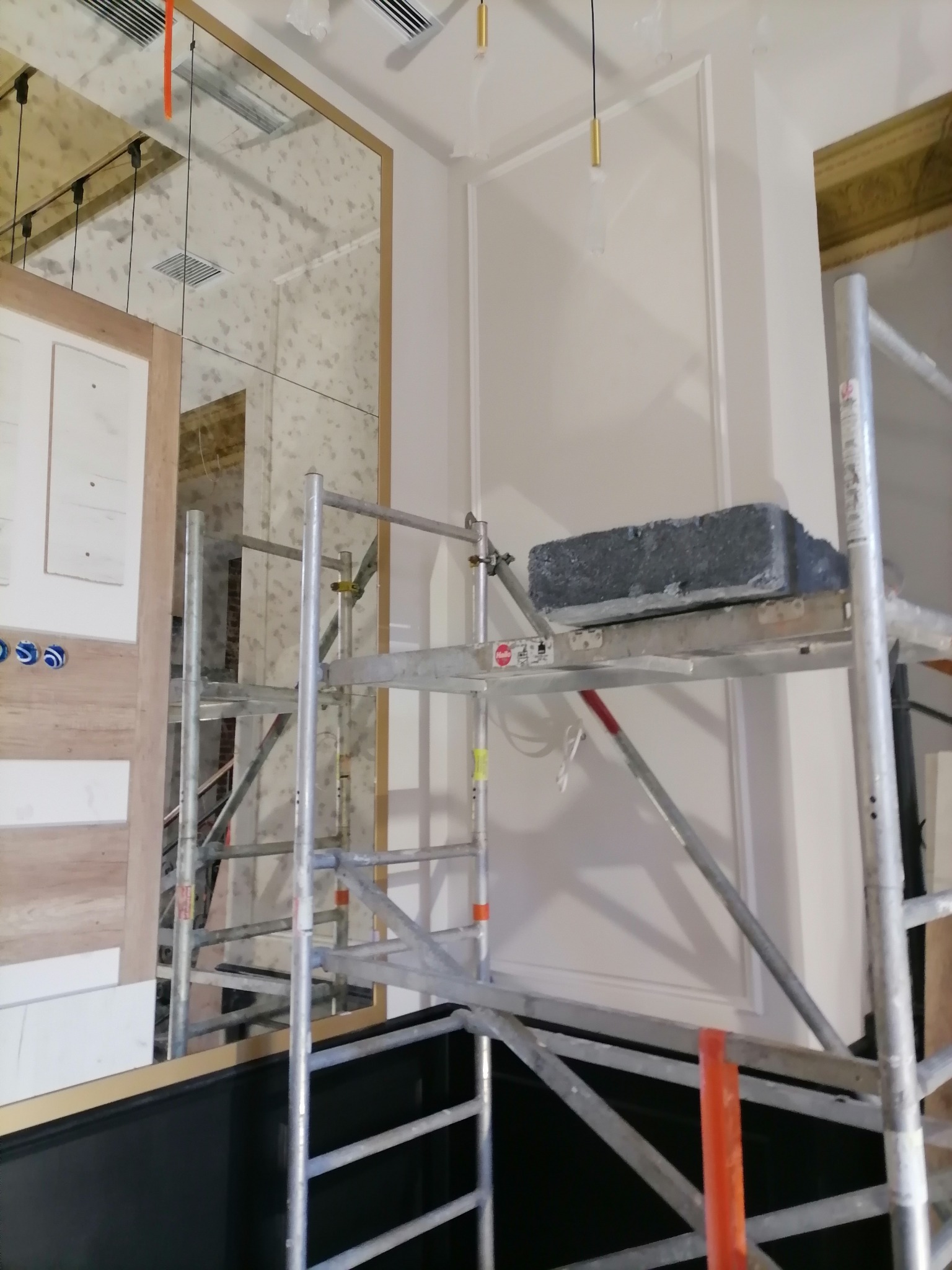 Wnętrze w trakcie prac wykończeniowych: widoczne rusztowanie z bloczkiem gipsowym, lustro z efektem postarzenia, listwy dekoracyjne na ścianie i fragment instalacji elektrycznej.