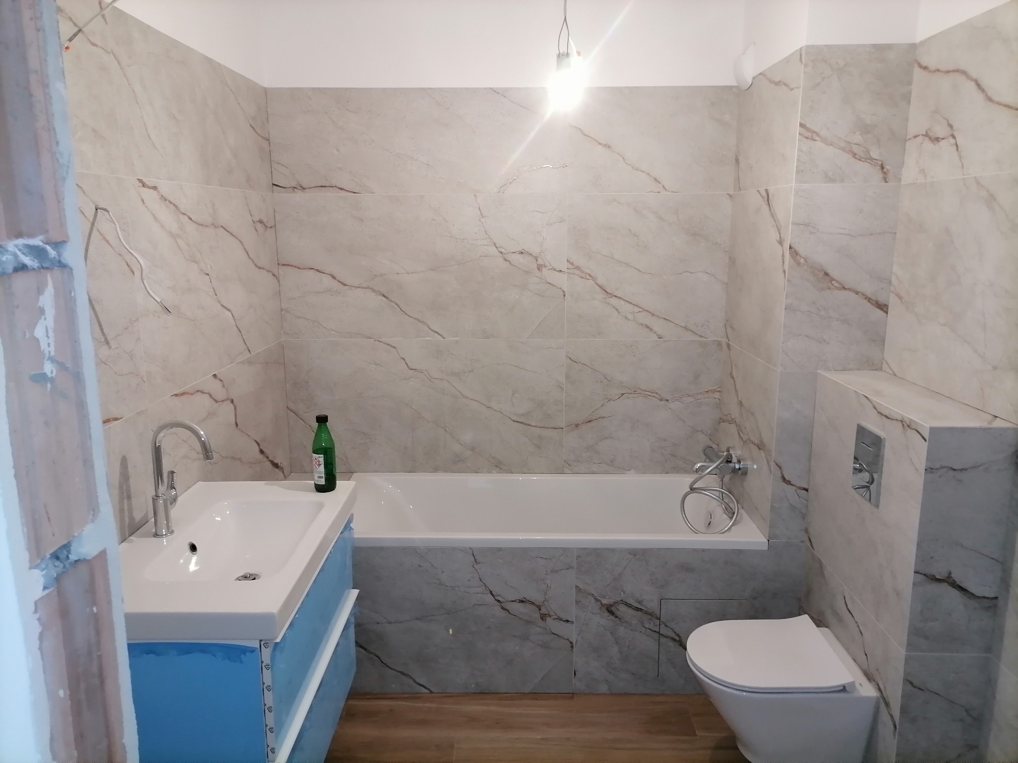 Wykończona łazienka z marmurowymi płytkami, białą wanną i umywalką z szafką zabezpieczoną folią, widok z perspektywy wejścia.