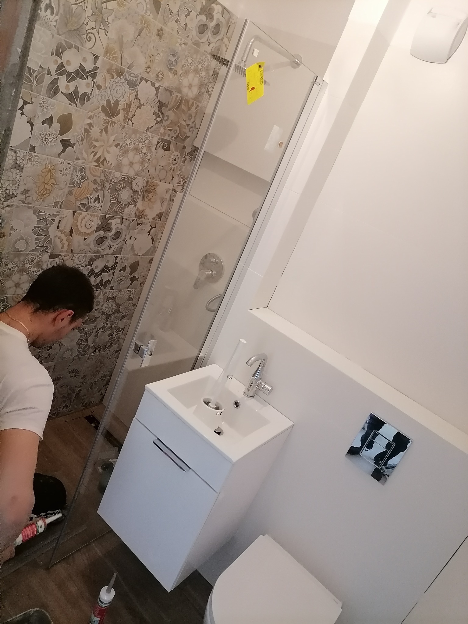 Montaż kabiny prysznicowej w łazience z wzorzystymi płytkami, widoczny hydraulik uszczelniający silikonem, biała umywalka z szafką i toaleta w tle.