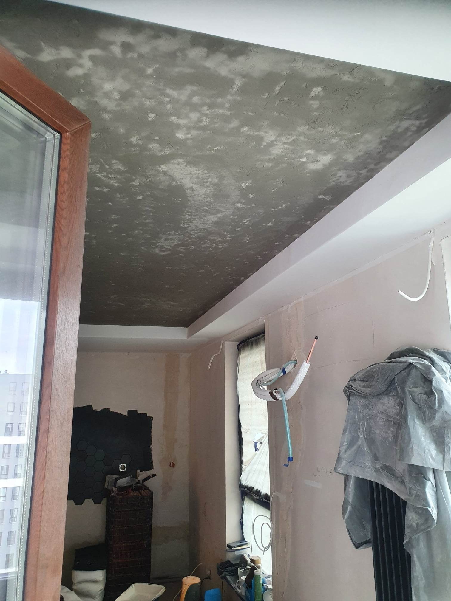Surowy betonowy sufit w trakcie wykańczania, widoczne przewody, okno z drewnianą ramą i fragment ściany z czarnymi heksagonalnymi płytkami, pomieszczenie w trakcie prac.