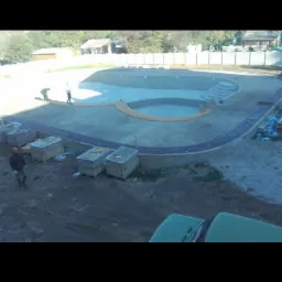 Budowa basenu z obrzeżem z kostki granitowej, widoczni pracownicy oraz palety z materiałem budowlanym.