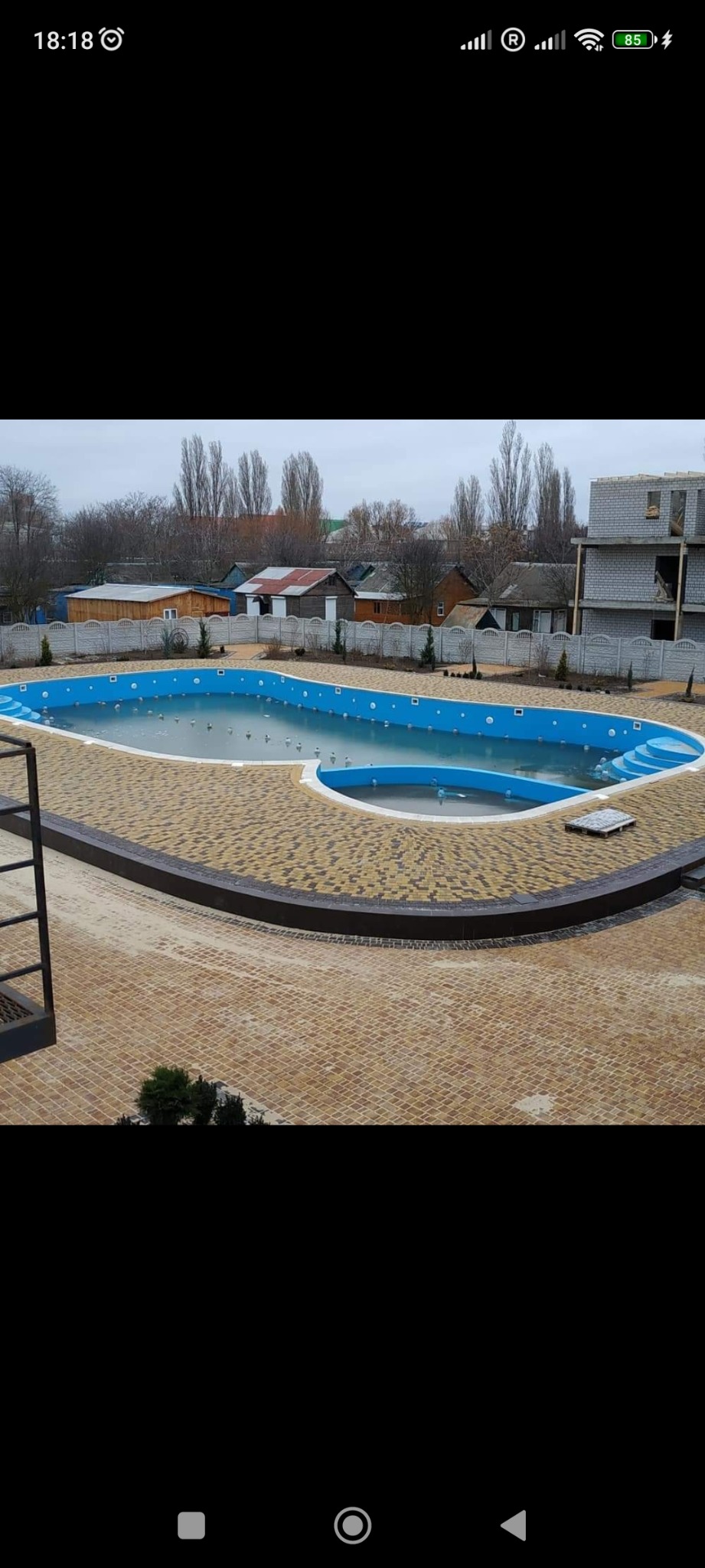 Nowoczesny basen zewnętrzny z brązową kostką brukową wokół, częściowo napełniony wodą, w tle budynki i drzewa.