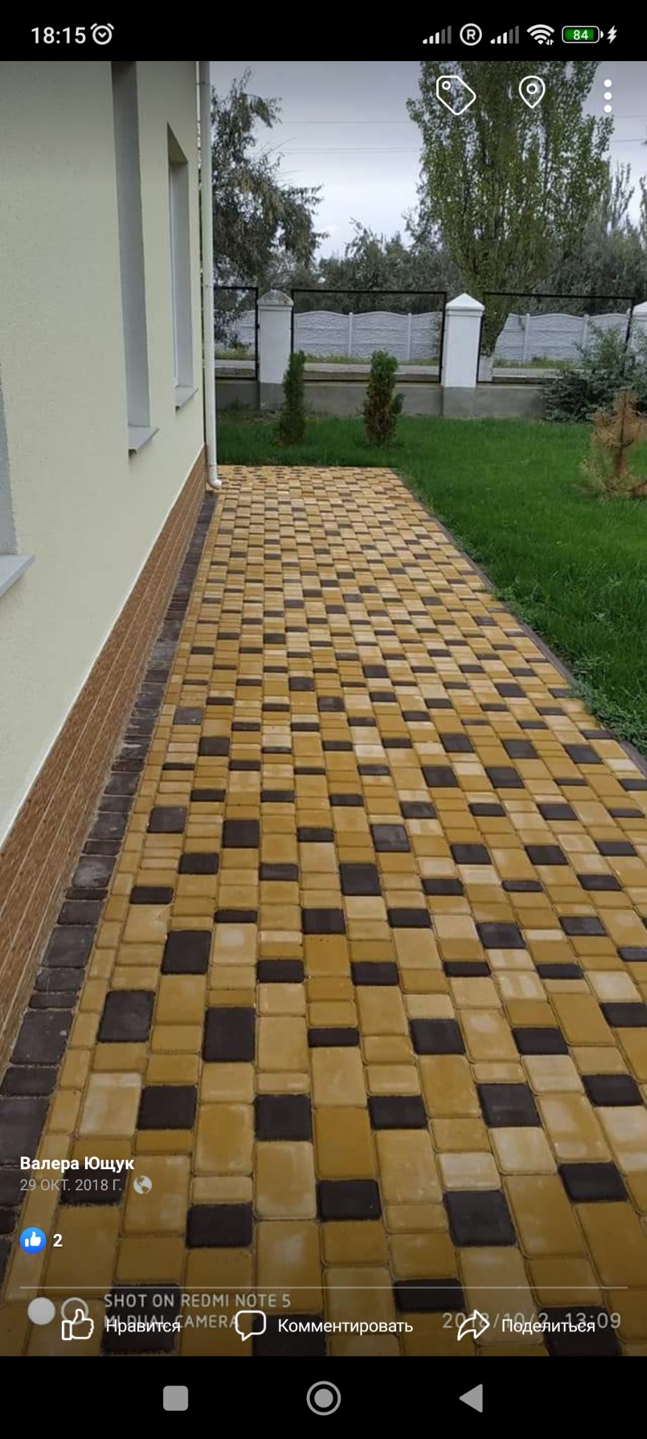 Ułożona ścieżka z kostki brukowej w kolorach żółtym i ciemnobrązowym, prowadząca wzdłuż jasnego budynku z brązową podmurówką, widok na trawnik i ogrodzenie w tle.