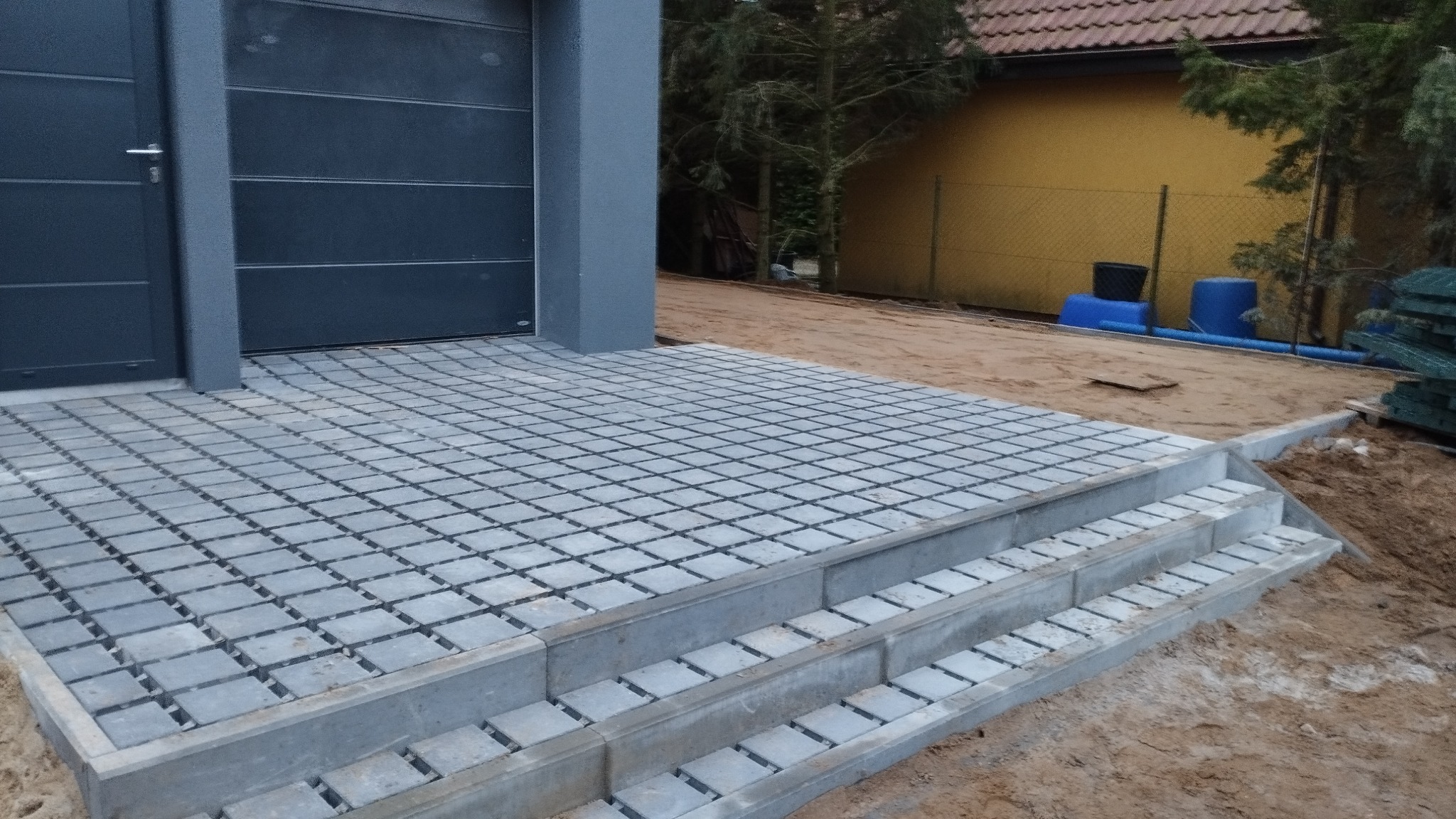 Szara kostka ażurowa ułożona przed garażem z trzema stopniami, widoczny teren przygotowany pod dalsze prace ziemne.