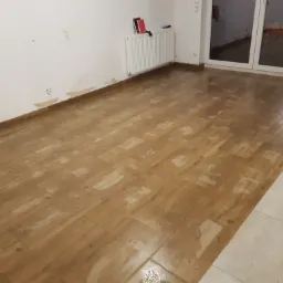 Parkiet ułożony w jodełkę, częściowo zabrudzony klejem, ściany przygotowane do odświeżenia, widoczny grzejnik i okno balkonowe.