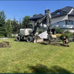 Biała koparka Terex wykonuje wykop w ogrodzie z wyznaczonym pomarańczową linią obrysem, mężczyzna nadzoruje prace, w tle nowoczesny dom z panoramicznymi oknami.