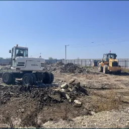 Biała koparka kołowa Terex i żółta ładowarka na placu budowy z gruzem i fragmentami betonu, widoczne ogrodzenie z blachy trapezowej i błękitne niebo.