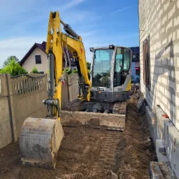 Żółta mini koparka Wacker Neuson ET58 na placu budowy, widoczny wykop wzdłuż fundamentu budynku z bloczków, ogrodzenie w tle.