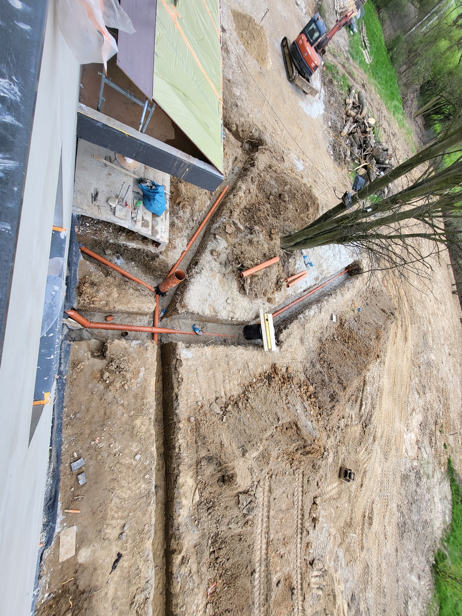 Widok z góry na wykop z ułożonymi pomarańczowymi rurami kanalizacyjnymi, częściowo zasypany ziemią, z widoczną koparką w tle i elementami konstrukcji budynku.