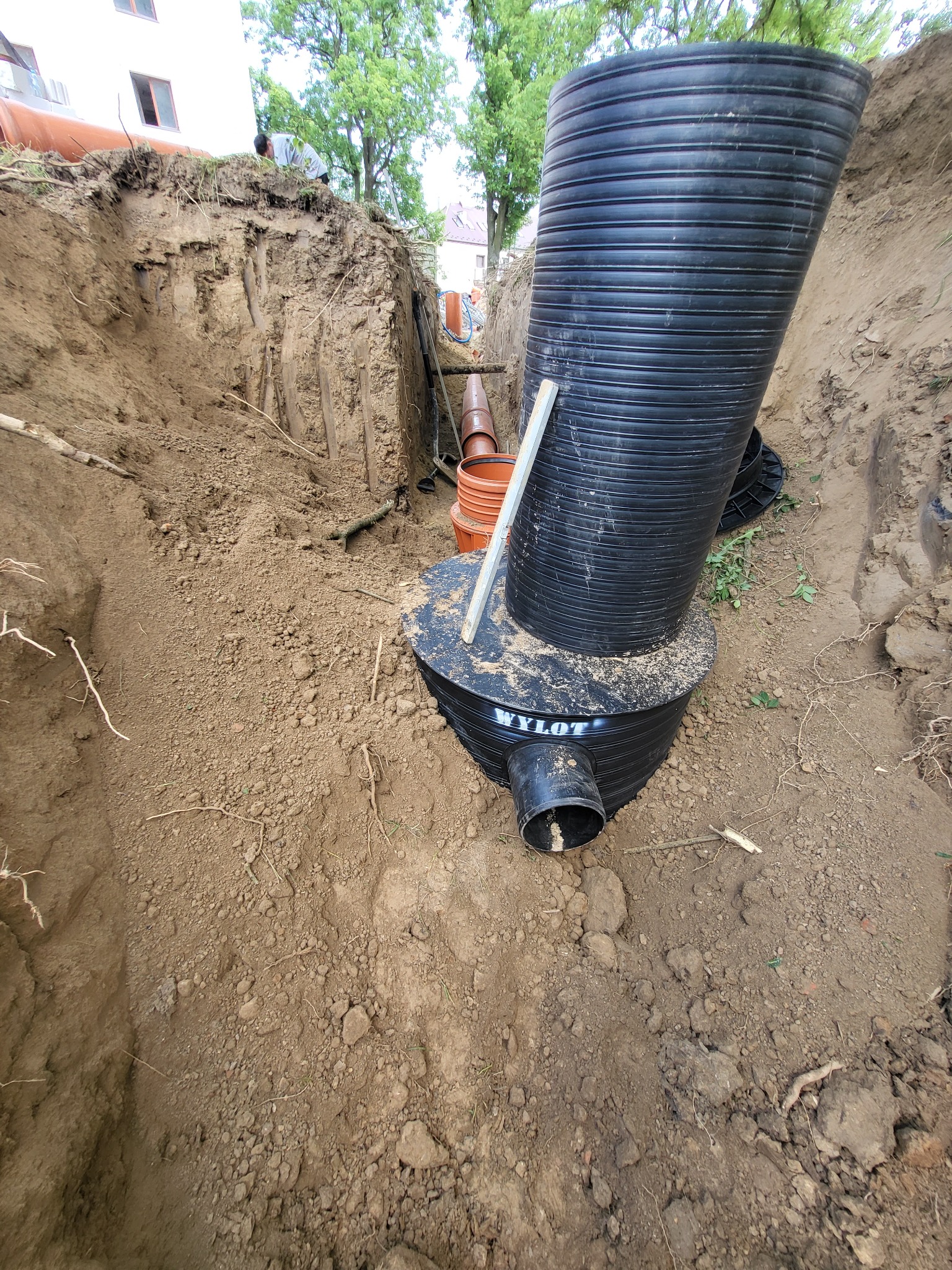 Głęboki wykop w ziemi z widoczną czarną rurą kanalizacyjną z tworzywa sztucznego, częściowo zamontowaną, z oznaczeniem producenta 'Wylot', oraz pomarańczowe rury kanalizacyjne w tle.
