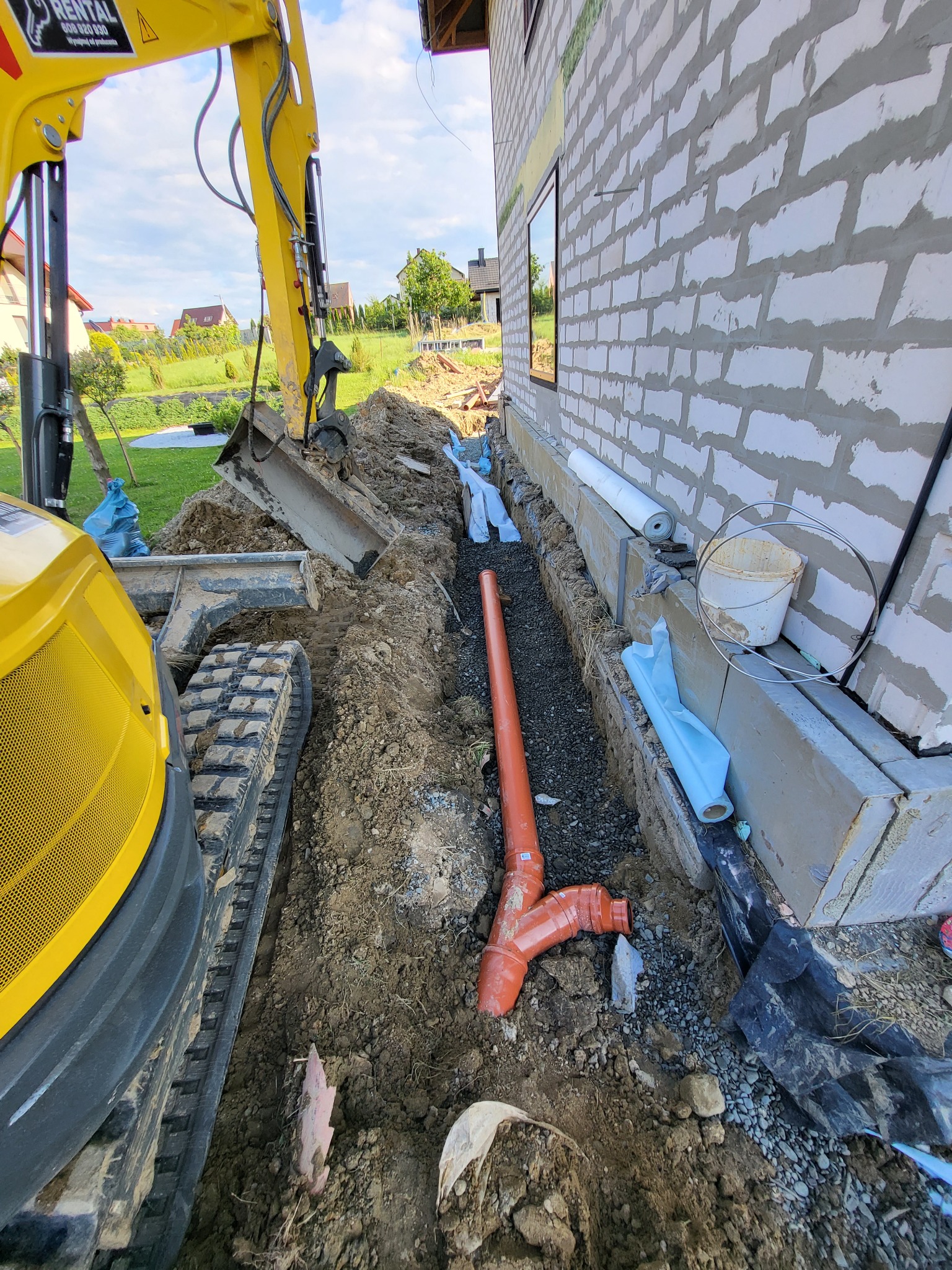 Żółta mini koparka obok wykopu pod fundamenty budynku z białego pustaka w Makowie Podhalańskim, widoczne pomarańczowe rury kanalizacyjne i przygotowane materiały izolacyjne.