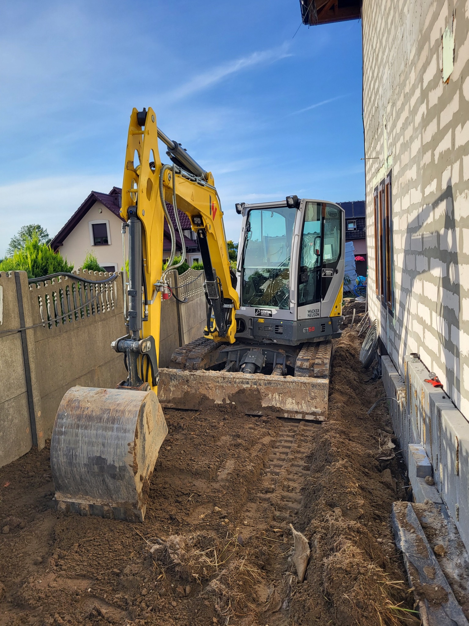 Żółta mini koparka Wacker Neuson ET58 na placu budowy, widoczny wykop wzdłuż fundamentu budynku z bloczków, ogrodzenie w tle.