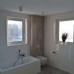 Nowoczesna łazienka z wanną, toaletą i drewnopodobną okładziną ściany za WC, z oknami z widokiem na zimowy krajobraz.