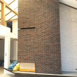 Wnętrze domu w trakcie prac wykończeniowych: ściana z cegły klinkierowej w odcieniach brązu, kontrastująca z fragmentem ściany pomalowanej na biało, widoczne worki z materiałami budowlanymi...