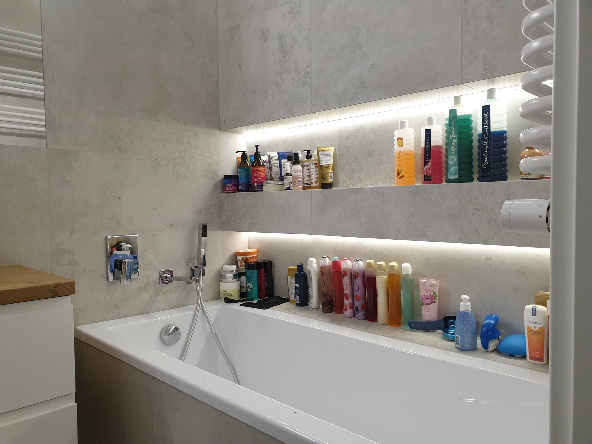 Nowoczesna łazienka z wanną, szarą płytką imitującą beton, oświetleniem LED w niszach i półkach z kosmetykami.