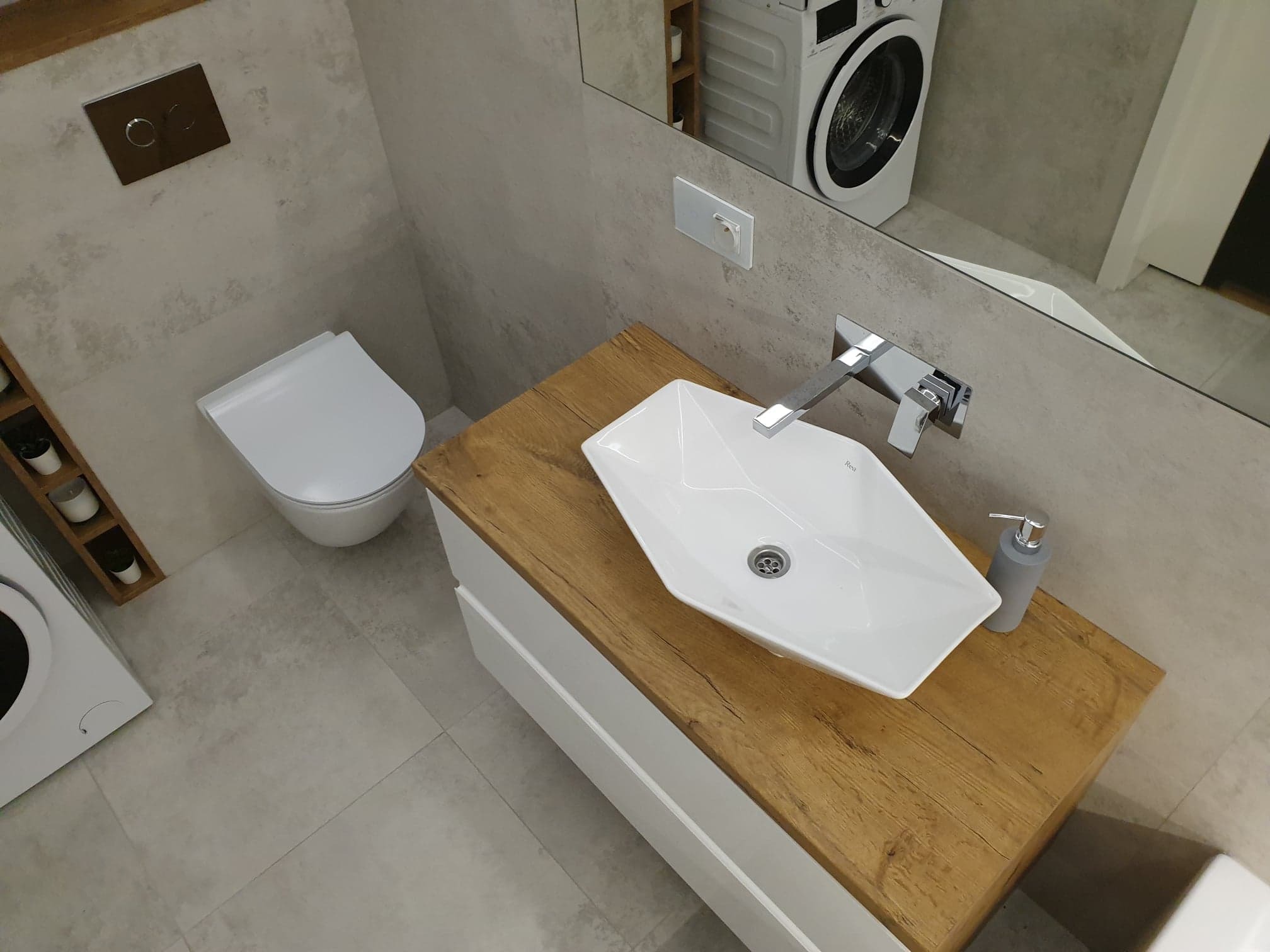 Nowoczesna łazienka z umywalką nablatową o geometrycznym kształcie, drewnianym blatem i szarą armaturą, widoczna toaleta i pralka w tle.