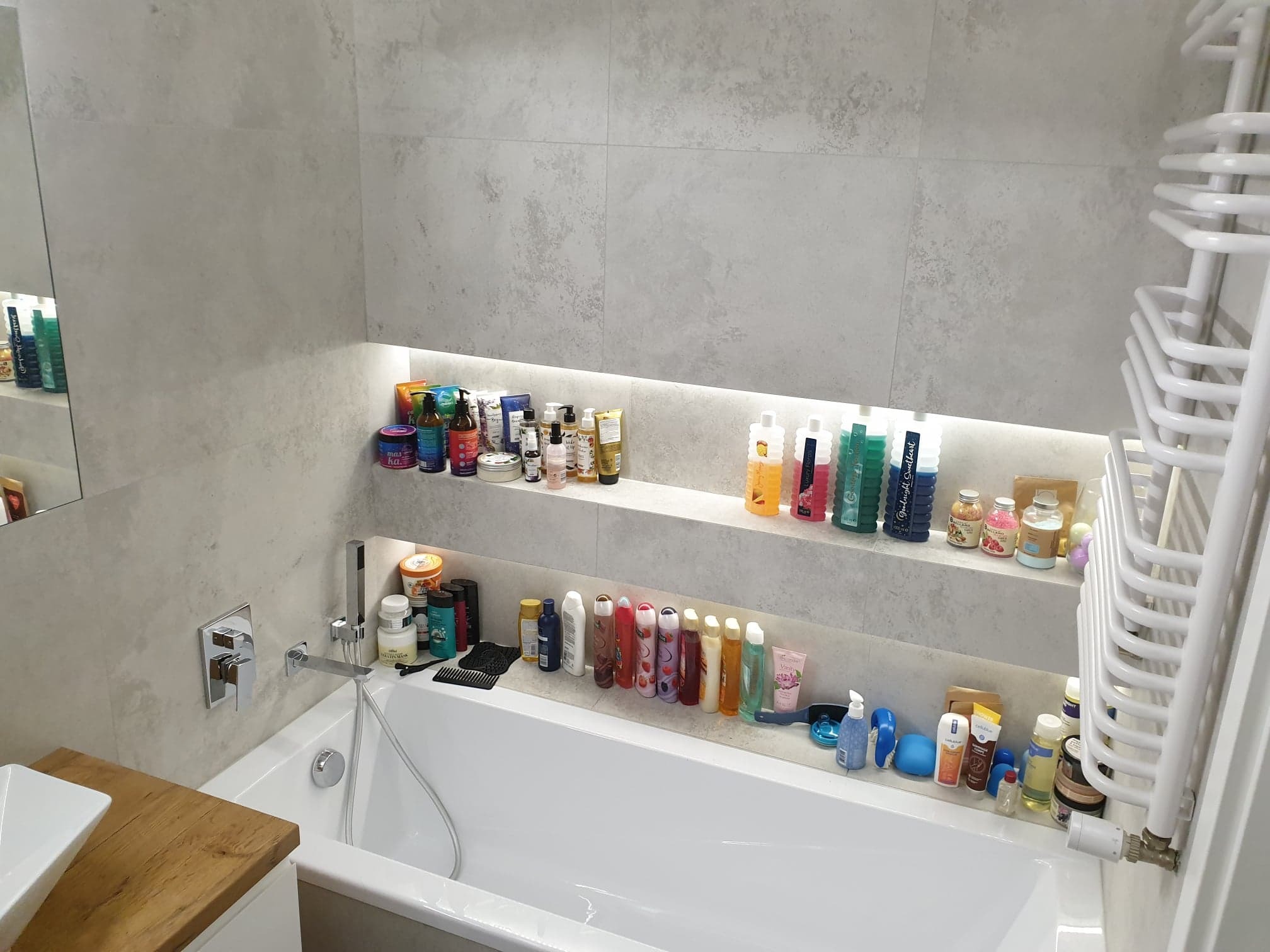 Wykończona łazienka z wanną, baterią podtynkową i dwiema wnękami w ścianie z szarych płytek, wypełnionymi kosmetykami i akcesoriami kąpielowymi, oświetlonymi od góry; grzejnik drabinkowy po prawej.