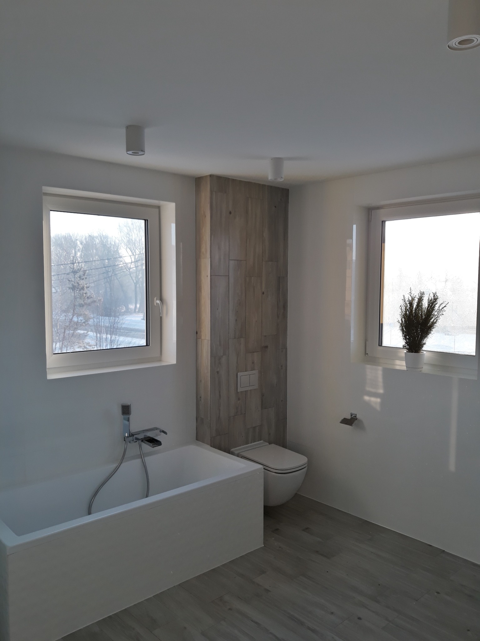 Nowoczesna łazienka z wanną, toaletą i drewnopodobną okładziną ściany za WC, z oknami z widokiem na zimowy krajobraz.