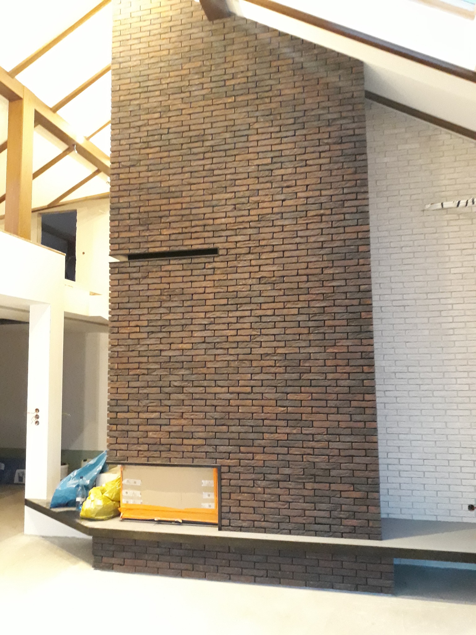 Wnętrze domu w trakcie prac wykończeniowych: ściana z cegły klinkierowej w odcieniach brązu, kontrastująca z fragmentem ściany pomalowanej na biało, widoczne worki z materiałami budowlanymi...