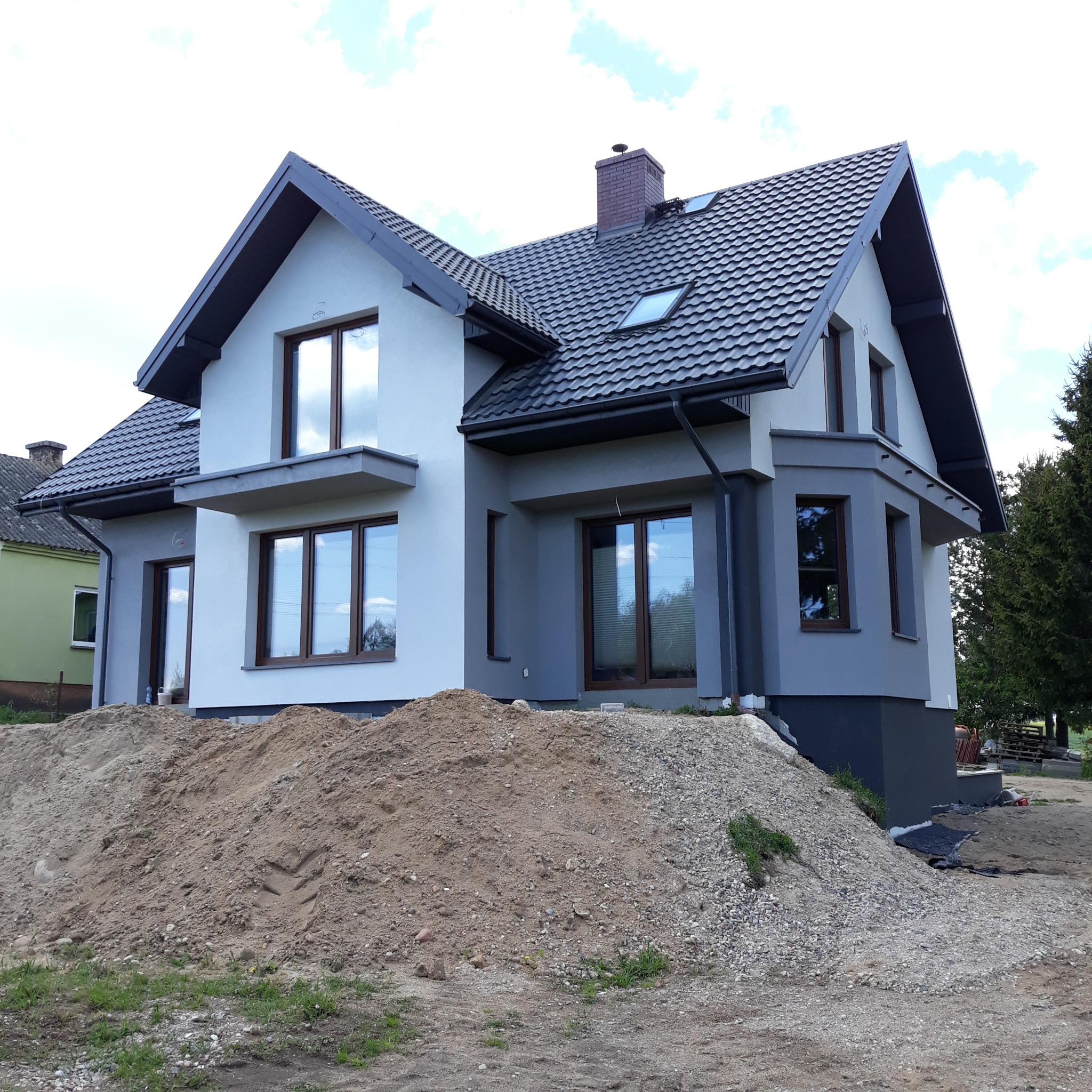 Nowoczesny dom jednorodzinny z szarą dachówką i elewacją w odcieniach szarości i bieli, widoczny stos piasku przed budynkiem, sugerujący prace wykończeniowe w toku.