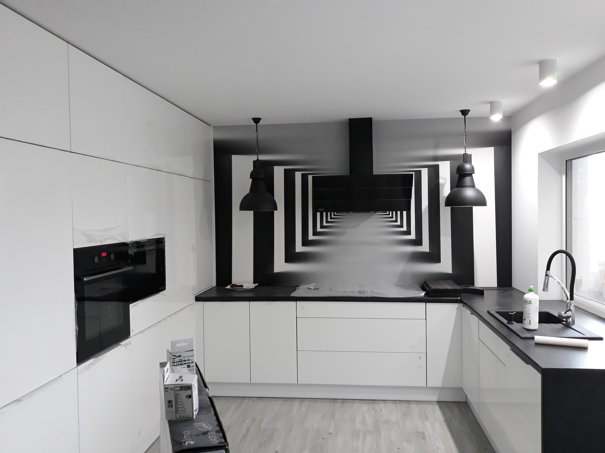 Nowoczesna kuchnia z białymi szafkami i czarnym blatem, ozdobiona fototapetą z motywem tunelu, widoczna drabina z akcesoriami montażowymi, lampy wiszące w stylu industrialnym.