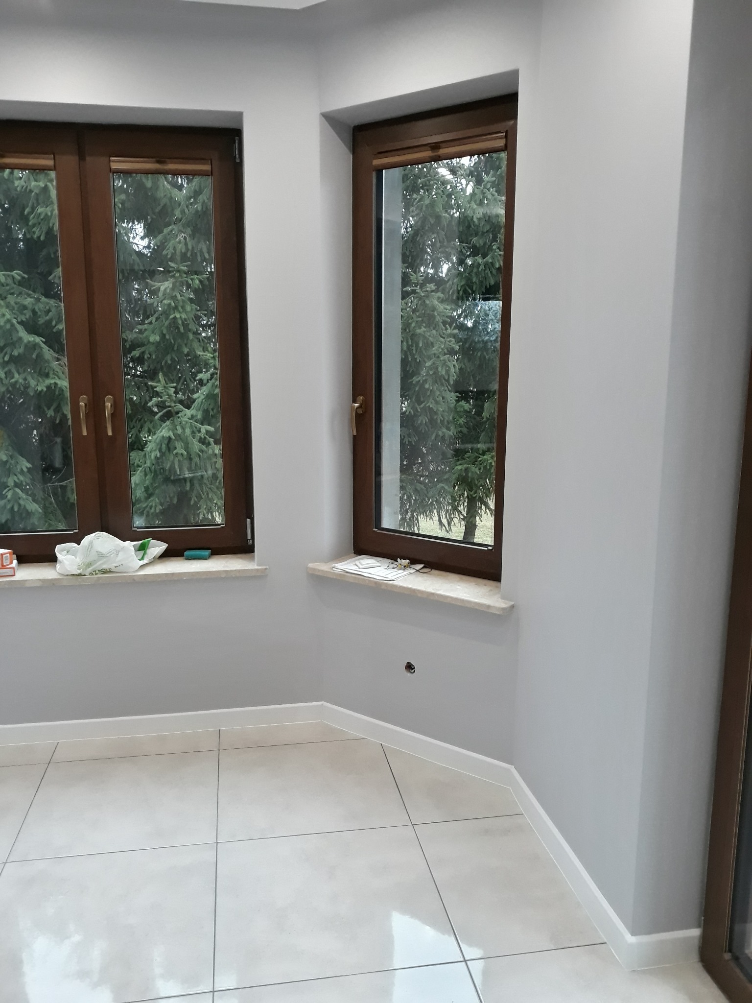 Pomalowane na szaro wnętrze pokoju z dwoma oknami w brązowych ramach, widok na zewnątrz na iglaki, jasne płytki podłogowe, białe listwy przypodłogowe.