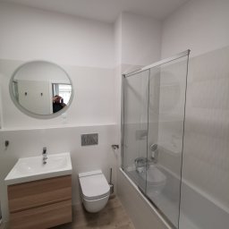 Usługi Remontowe - Nowoczesna, jasna łazienka w Łodzi po remoncie: okrągłe lustro z podświetleniem nad drewnianą szafką umywalkową, podwieszana toaleta i wanna z parawanem nawannowym.