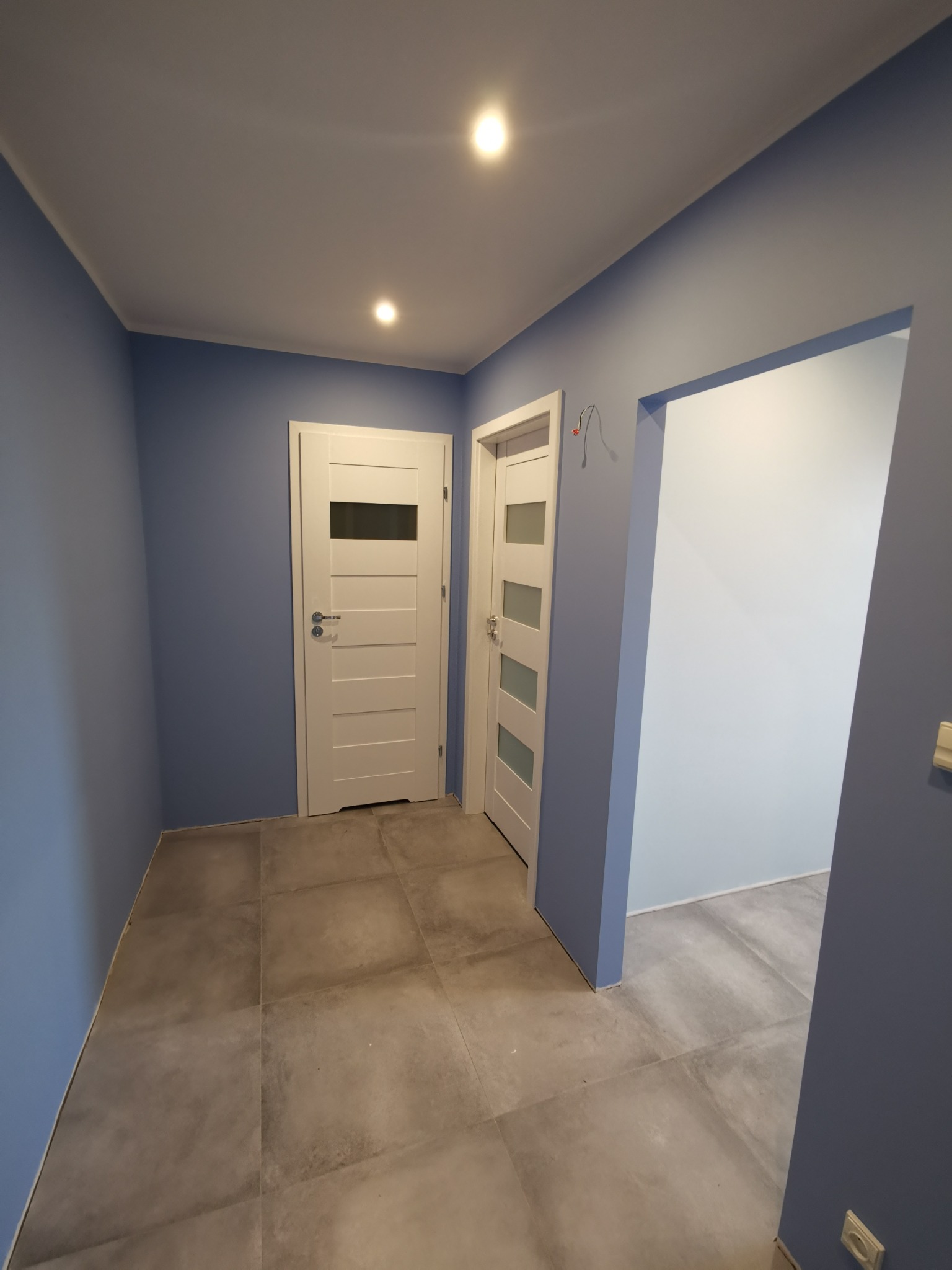 Korytarz po remoncie z niebieskimi ścianami, szarą podłogą z płytek i dwoma białymi drzwiami z przeszkleniami.