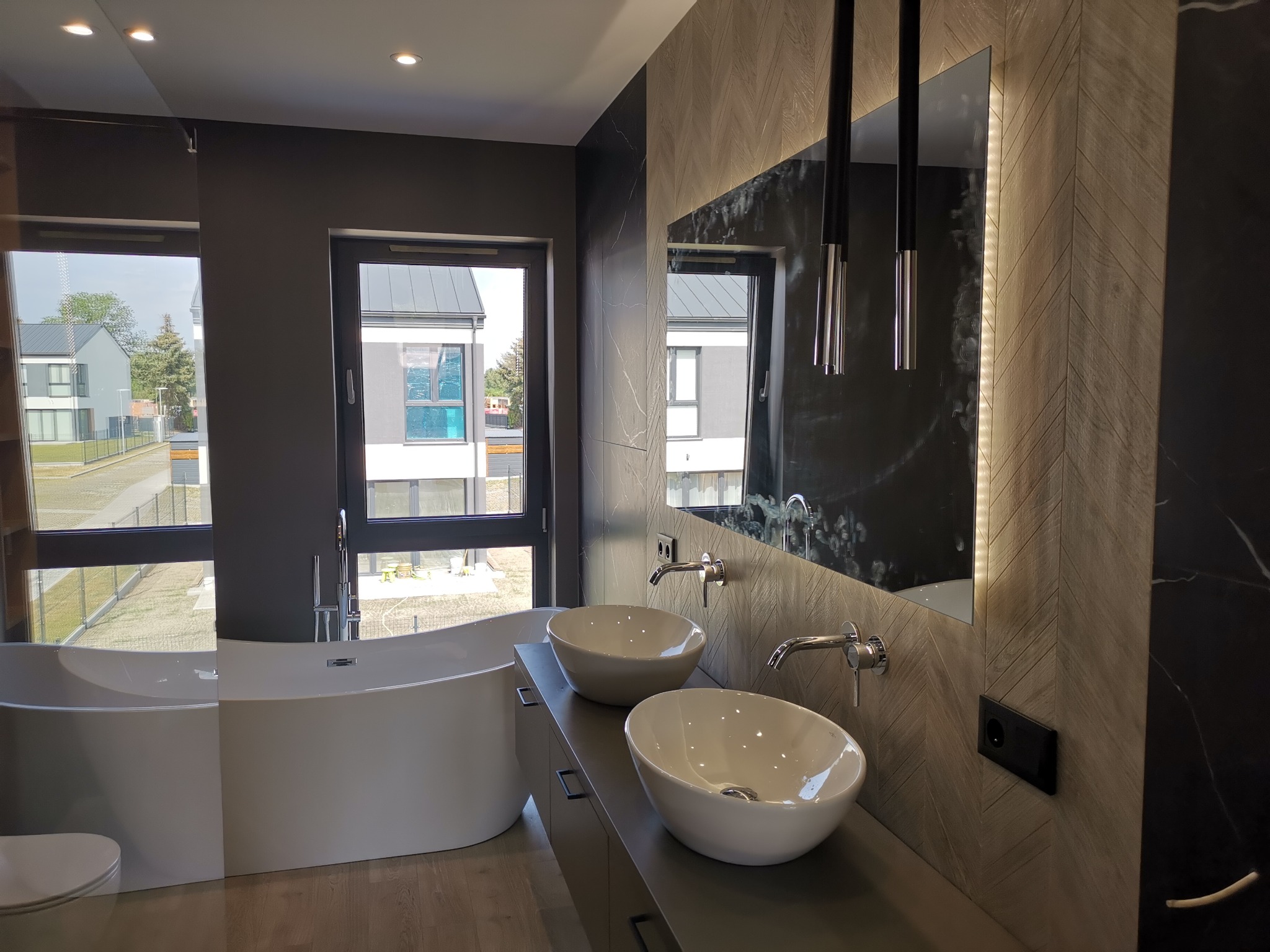 Nowoczesna łazienka z wolnostojącą wanną, dwiema umywalkami nablatowymi i lustrem z podświetleniem LED, wykończona płytkami imitującymi drewno ułożonymi w jodełkę.