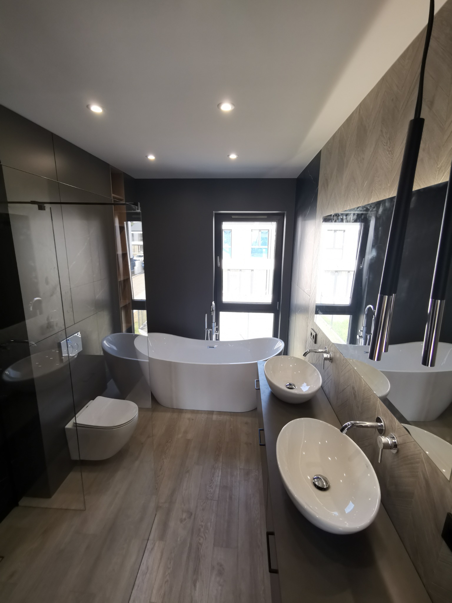 Nowoczesna łazienka z wolnostojącą wanną, dwiema owalnymi umywalkami nablatowymi, szarą ścianą i drewnopodobną podłogą, oświetleniem punktowym i dużym lustrem.