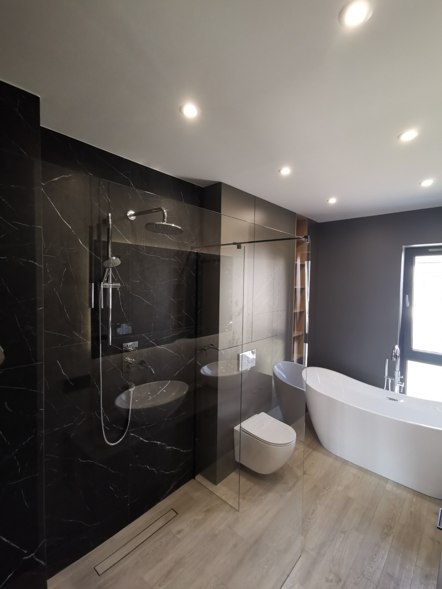 Nowoczesna łazienka z czarnymi marmurowymi płytkami pod prysznicem, wolnostojącą białą wanną i podwieszaną toaletą, oświetlona punktowymi lampami sufitowymi.