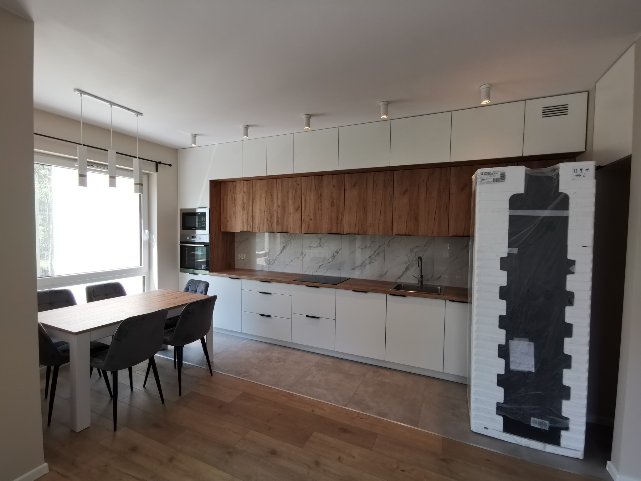 Nowoczesna kuchnia z białymi i drewnianymi szafkami, marmurowym backsplashiem, stołem z szarymi krzesłami oraz zapakowaną lodówką czekającą na montaż.