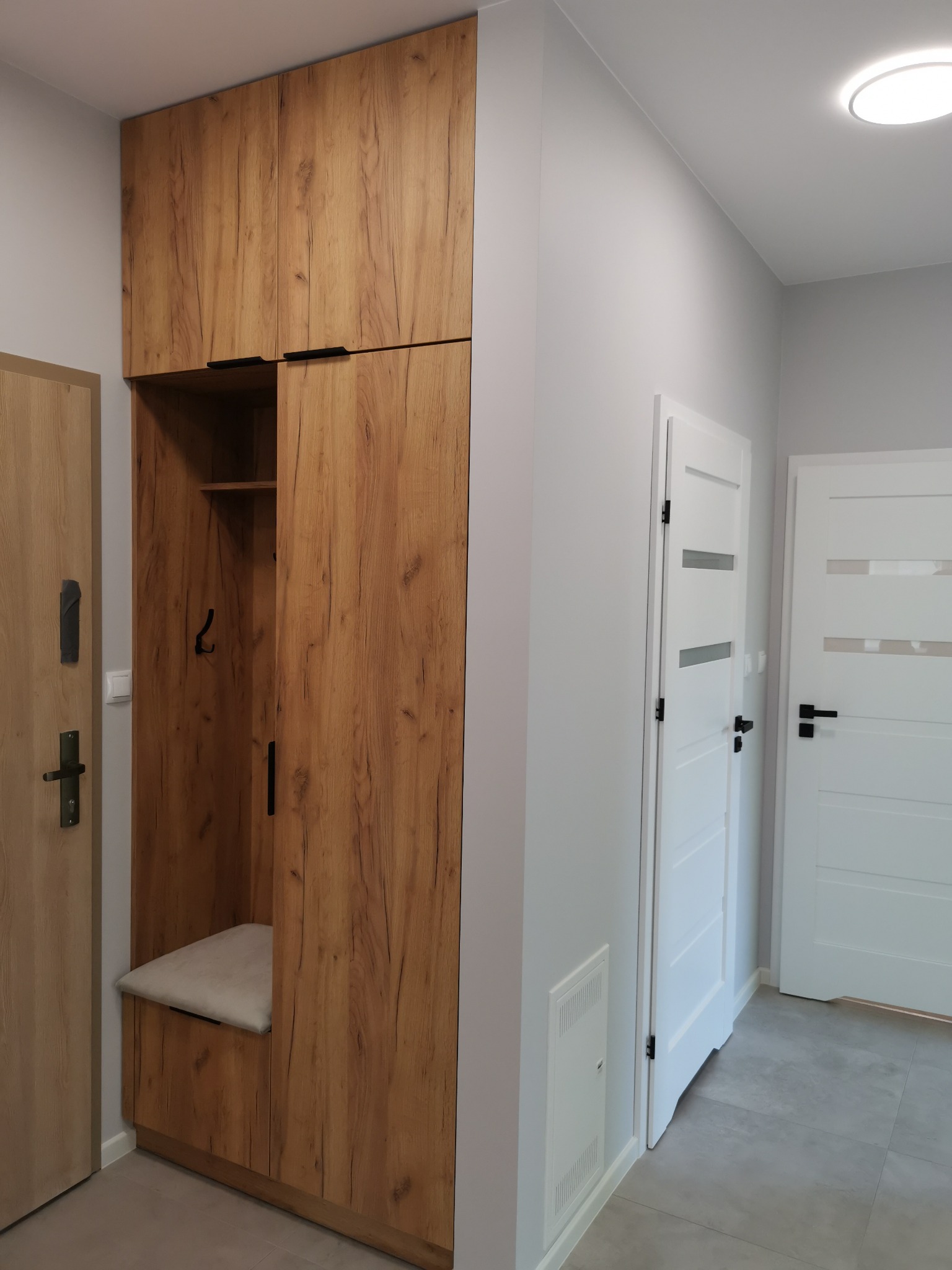 Wnętrze przedpokoju z zabudową meblową z siedziskiem i wieszakami, drewnopodobna okleina, szare ściany, białe drzwi wewnętrzne.