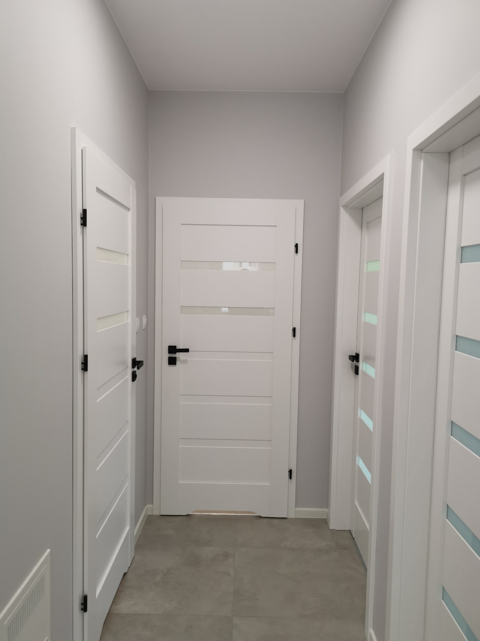 Wnętrze korytarza z trzema białymi drzwiami z przeszkleniami i czarnymi klamkami, szare ściany i podłoga z płytek w odcieniach szarości.