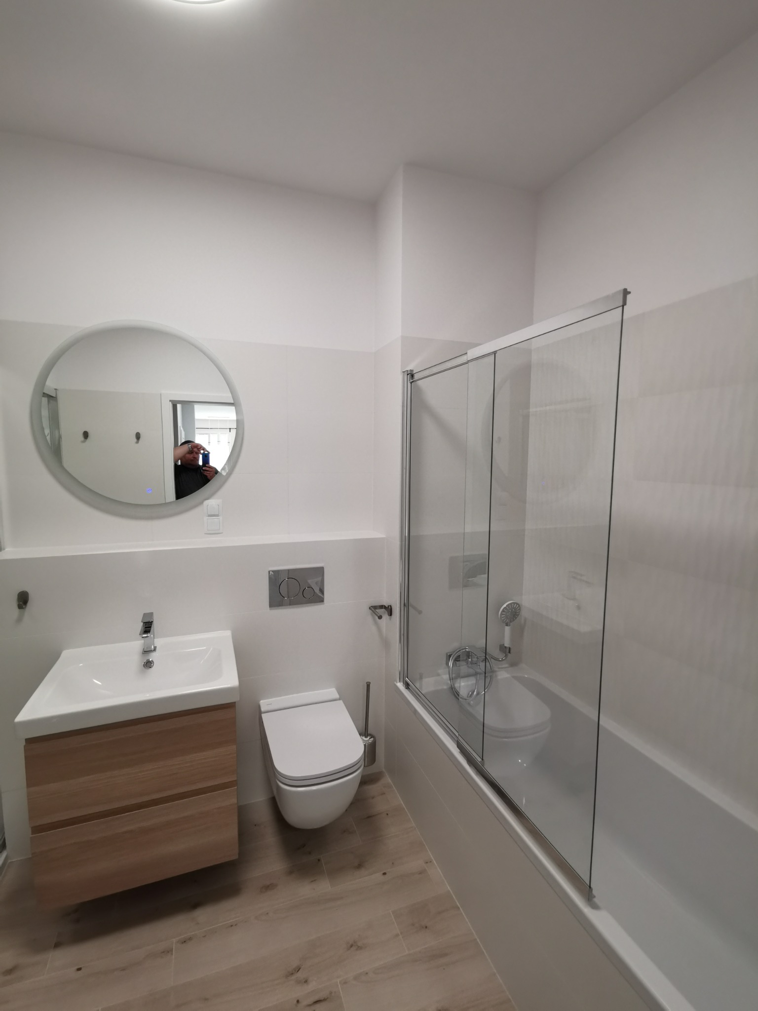 Nowoczesna, jasna łazienka w Łodzi po remoncie: okrągłe lustro z podświetleniem nad drewnianą szafką umywalkową, podwieszana toaleta i wanna z parawanem nawannowym.