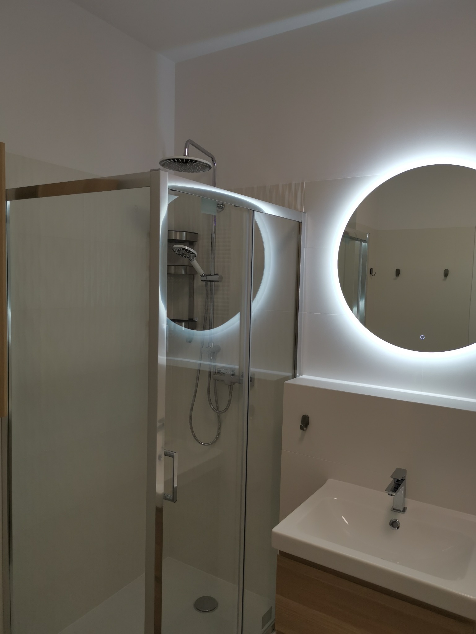 Nowoczesna łazienka z kabiną prysznicową ze szklanymi drzwiami i okrągłym lustrem z podświetleniem LED, umywalka nablatowa na drewnianej szafce, białe płytki na ścianach.