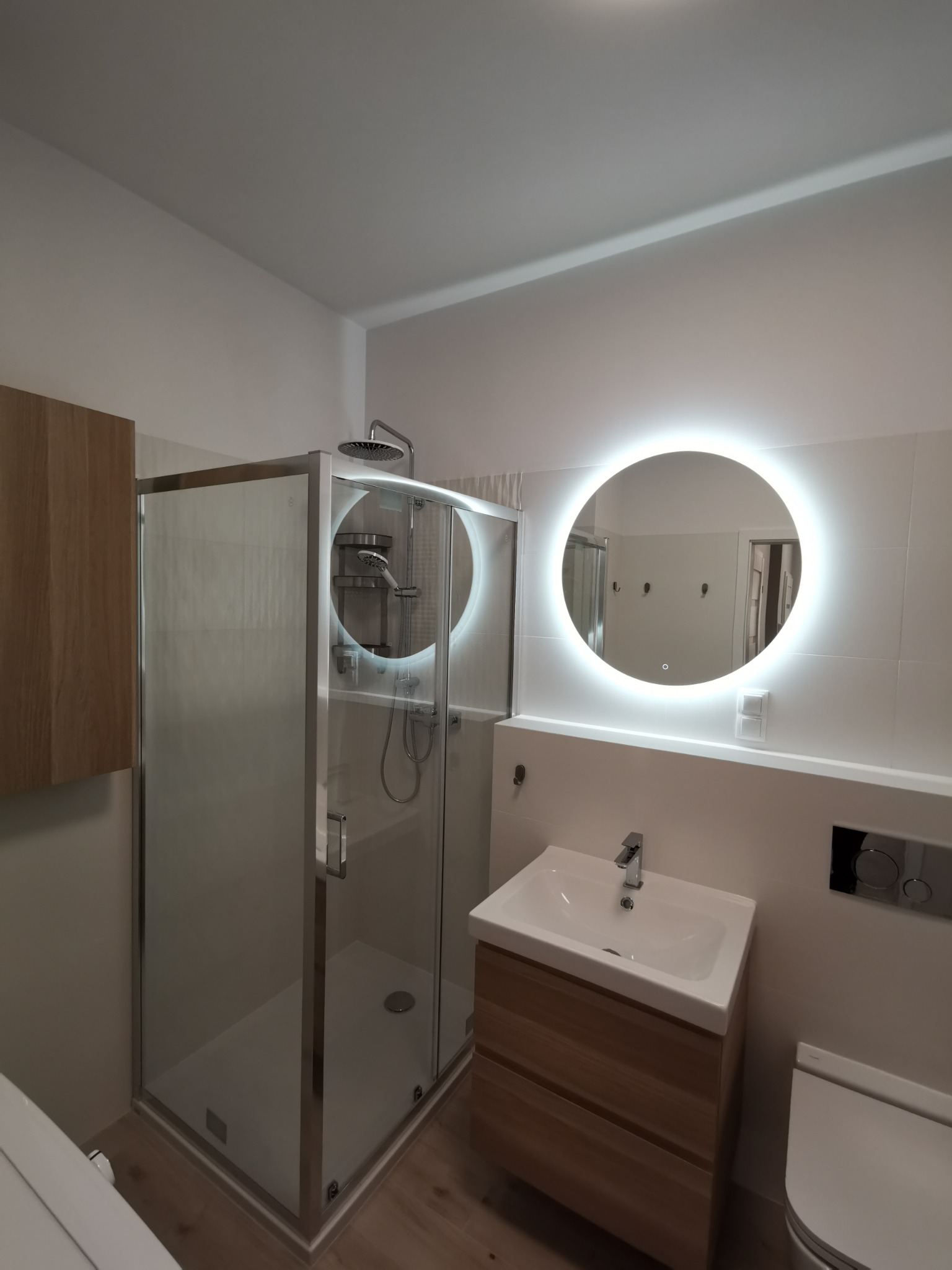 Nowoczesna łazienka z kabiną prysznicową ze srebrnymi ramami, umywalką z szafką z jasnego drewna i okrągłym lustrem z podświetleniem LED.