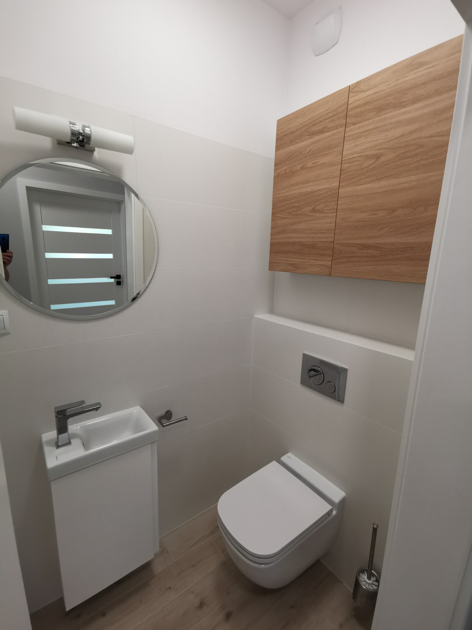 Nowoczesna, mała łazienka z podwieszaną toaletą, umywalką na szafce, okrągłym lustrem z oświetleniem i szafką z drewnopodobnym frontem.