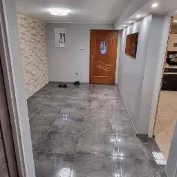 Wyremontowane wnętrze z szarymi płytkami podłogowymi z marmurkowym wzorem, drewnianymi drzwiami i fragmentem ściany wykończonej dekoracyjną cegłą, oświetlone punktowymi lampami sufitowymi.