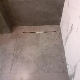 Szare płytki podłogowe z odpływem liniowym w rogu łazienki, białe płytki marmurowe na ścianie.