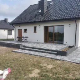 Nowoczesny taras z kostki brukowej w odcieniach szarości, prowadzący do białego domu z ciemną dachówką, widok z poziomu trawnika.