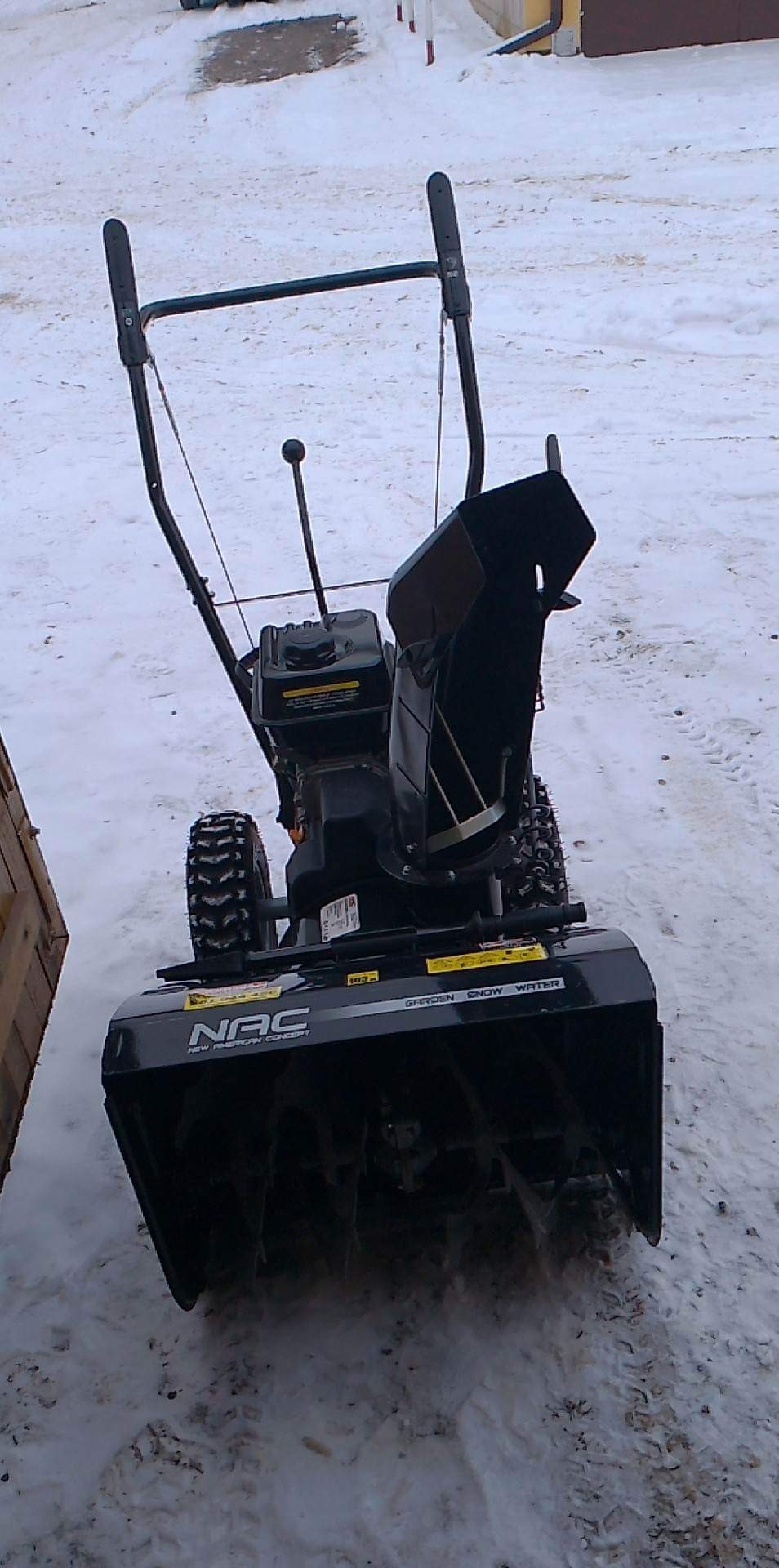 Czarno-szara odśnieżarka NAC stoi na śniegu, widoczne ślady opon w tle i fragment drewnianej skrzyni z lewej strony.