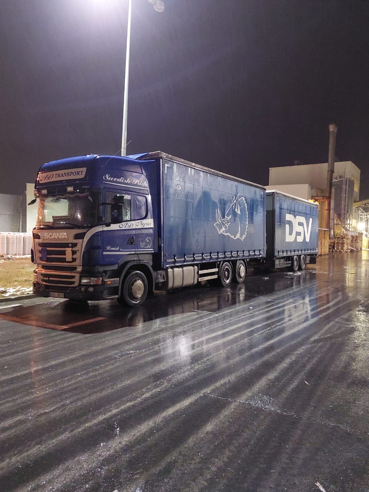 Niebieska ciężarówka Scania z naczepą DSV, zaparkowana na mokrym, oświetlonym placu w nocy, z widocznym logo firmy transportowej i napisem 'Swedish Pride' na kabinie.